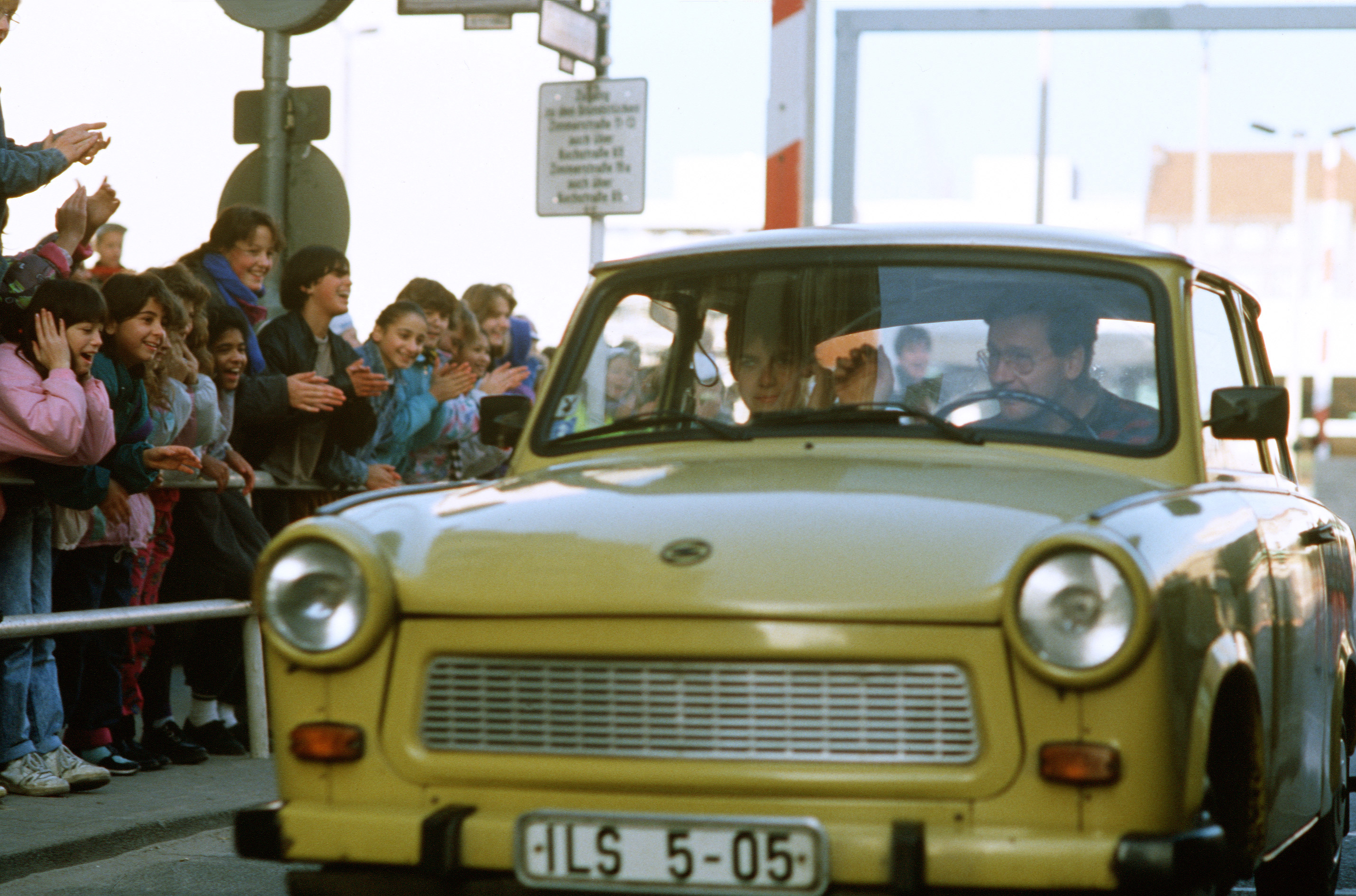 West German children applaud as an East German couple drive through Checkpoint Charlie and take advantage of relaxed travel restrictions to visit West Germany.
