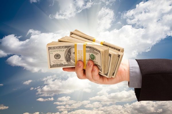 Male Hand Holding Stack of Cash Over Dramatic Clouds and Sky with Sun Rays.