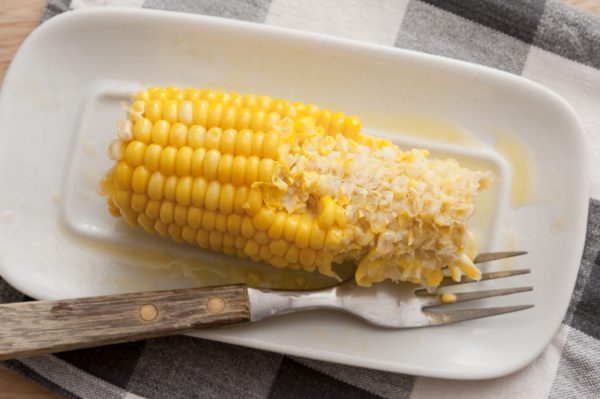 Half eaten cob of sweet corn with a fork and melted butter on a white plate viewed from above